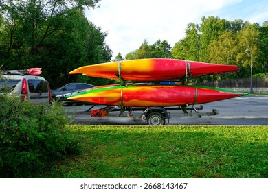 Side view small trailer is loaded with several colorful kayaks and canoes in preparation for a fun day of summer water recreation. The trailer is parked near a lake in the Park.