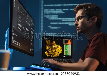 Side view of smart programmer man looking at big monitor checking id-address and debugging system, writing html code, sitting at table late at night on zoomed digital background