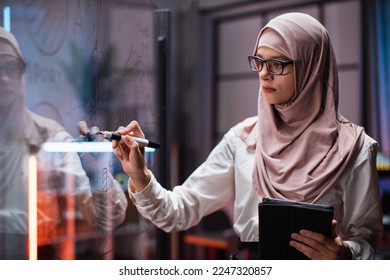 Side view of smart young businesswoman in hijab, brainstorming near office glass board, using tablet. Confident female employee writing on glass wall, developing business project at workplace. - Powered by Shutterstock