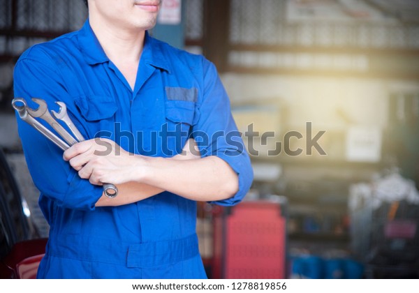 Side view\
smile professional asian technician engineer man in blue uniform is\
holding wrench at outdoor garage with yellow light using for\
automobile industrial copy space or report\

