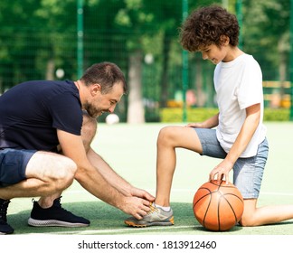 Side View Of Smiling Adult Dad Helping His Son To Tie Shoelaces, Kid Playing On Basketball Field