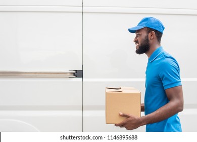 side view of smiling african american delivery man carrying cardboard box 