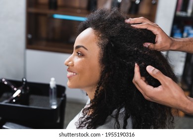 Side view of smiling african american client sitting near hairstylist touching hair