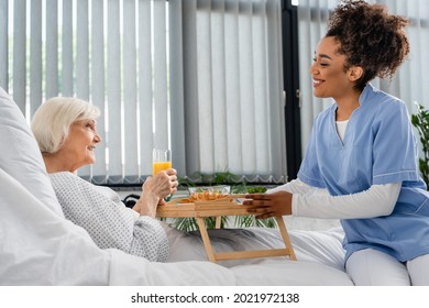 Side View Of Smiling African American Nurse Holding Tray With Food Near Patient With Orange Juice In Hospital Ward