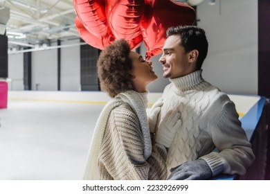 Side view of smiling african american woman looking at boyfriend near heart shaped balloons on ice rink  - Shutterstock ID 2232926999