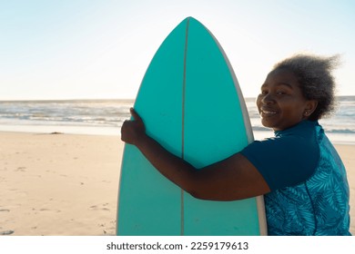 Side view of smiling african american senior woman with surfboard standing at beach over sea and sky. Copy space, water sports, recreation, retirement, unaltered, sunset, vacation, enjoyment, nature. - Powered by Shutterstock