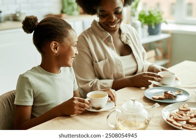 Side view of smiling African American little daughter cheerfully talking to young mother, while having tea with sweet treat together at family gathering at kitchen table - Powered by Shutterstock