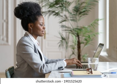 Side View Of Smiling Afro Woman In Blazer Sitting On Desk, Remote Working At Laptop At Home Office, Studying. Confident Black Employee Female Talking In Video Chat At Computer. Distance Job.