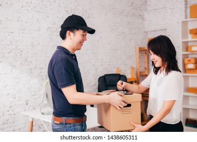 Side View Of Smiling Asian Female Signing Tablet While Taking Carton Box From Happy Delivery Man In Office