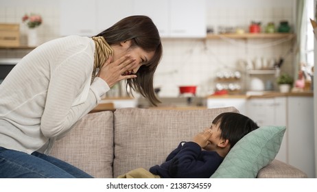 Side View Of Smiling Asian Mother And Son Making Silly Face To Each Other. The Boy Is Lying On The Sofa While Playing With His Mom At Home