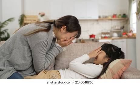 Side View Of Smiling Asian Mother And Daughter Making Silly Face And Cove The Face First To Each Other In Living Room. The Girl Is Lying On The Sofa While Playing With Her Mom At Home