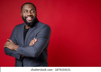 Side View Of Smiling Attractive Young African American Business Man 20s In Classic Jacket Suit Standing Holding Hands Crossed Looking Aside Isolated On Bright Red Color Background Studio Portrait