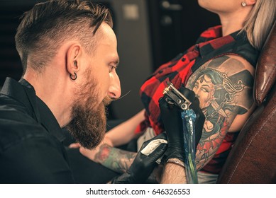 Side View Of Smiling Bearded Man Doing Tattoo On Arm Of Young Woman. She Leaning At Table