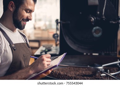 Side view smiling bearded man writing information about roasting beans in special equipment. Job concept
