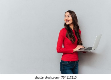 Side View Of Smiling Brunette Woman In Red Blouse Holding Laptop Computer And Looking Back Over Gray Background