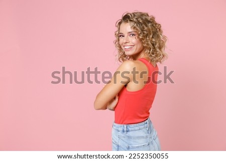Side view of smiling cheerful attractive young blonde woman 20s wearing basic casual tank top standing holding hands crossed looking camera isolated on pastel pink colour background, studio portrait