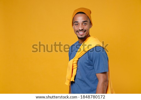 Side view of smiling cheerful funny handsome young african american man 20s wearing basic casual blue t-shirt hat standing looking camera isolated on bright yellow colour background, studio portrait