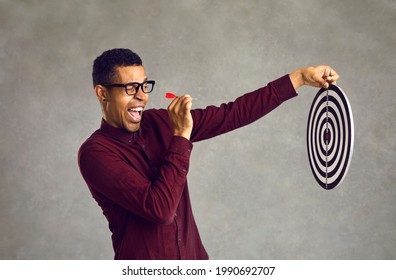 Side view smiling confident young black man aiming red dart at shooting target determined to throw it and hit bullseye. Using chance, accepting challenge, achieving business goal and objective concept