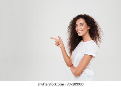 Side view of smiling curly woman pointing away and looking at the camera over gray background