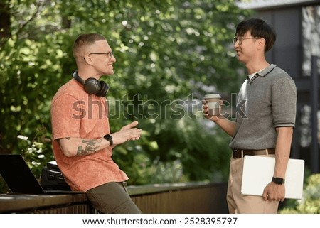 Side view of smiling IT developer with headphones chatting with cheerful partner holding laptop and reusable cup of coffee during meeting in park, copy space