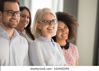 Side view of smiling diverse employees stand in row together posing for group picture in office, happy confident multiethnic team look at camera making photo, showing unity. Teamwork concept
