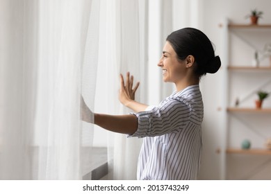 Side view smiling dreamy Indian woman opening curtains in early morning, looking out window in distance, thinking and dreaming, happy beautiful young female feeling positive, starting new day