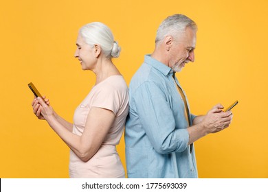 Side view of smiling elderly gray-haired couple woman man in casual clothes isolated on yellow background studio. People lifestyle concept. Mock up copy space. Using mobile phone, typing sms message