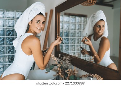 Side view of smiling ethnic female in white bodysuit and towel on head looking at camera during makeup session after morning shower in bathroom