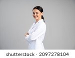 Side view of the smiling female doctor in lab coat with arms crossed looking away and posing against grey background. Medicine concept 
