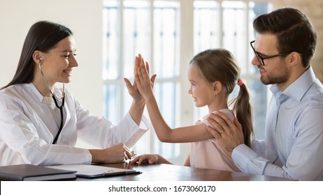 Side View Smiling Female Doctor Nurse Giving High Five To Little Preschool Girl Sitting On Daddy's Lap. Pleasant General Practitioner Getting Acquainted With Small Patient At Checkup In Modern Clinic.