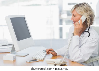 Side View Of A Smiling Female Doctor Using Phone And Computer At Desk In Medical Office