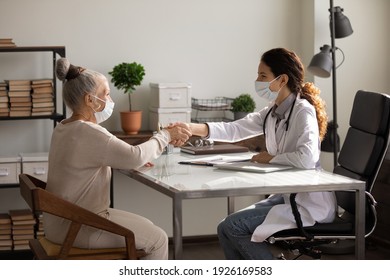 Side View Smiling Female Doctor Wearing Face Mask And Coat With Mature Patient Shaking Hands, Sitting At Table In Office, Greeting, Older Woman Making Health Insurance Deal, Successful Checkup