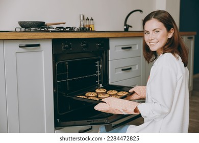 Side view smiling happy fun cheerful young housewife woman wear casual clothes baking chocolate cookies put into oven eat breakfast cooking food in light kitchen at home alone. Healthy diet concept - Powered by Shutterstock