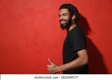 Side View Of Smiling Happy Young African American Man With Dreadlocks 20s Wearing Black Casual T-shirt Posing Showing Thumb Up Looking Camera Isolated On Bright Red Color Background Studio Portrait