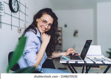 Side view of smiling long haired female in glasses in striped blue shirt sitting at laptop in workplace at home - Shutterstock ID 1548670535