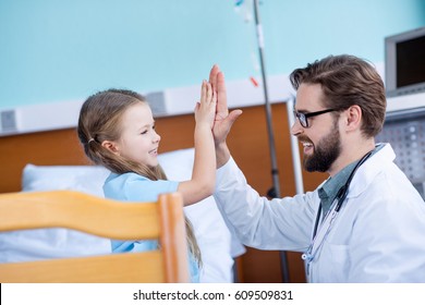 Side View Of Smiling Male Doctor And Cute Little Girl Giving High Five In Hospital