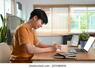 Side View Smiling Man Sitting In Front Of Laptop Computer And Making Notes On Notebook.