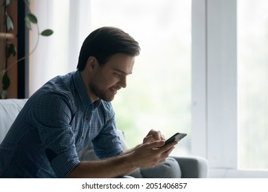 Side View Smiling Man Using Smartphone, Sitting On Cozy Couch At Home, Happy Young Male Looking At Phone Screen, Chatting Online, Typing Writing Message In Social Network, Shopping, Surfing Internet