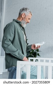 Side View Of Smiling Mature Man Holding Baby Bottle Near Crib