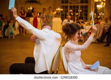 Side View Of The Smiling Newlyweds Who Are Sitting Back To Back On The Chairs In The Restaurant And Playing The Game