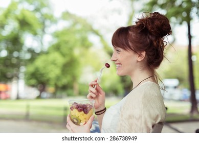 Side View of a Smiling Pretty Young Woman at the Park, Eating Fresh Fruit Salad on a Plastic Container While Looking Into Distance. - Powered by Shutterstock