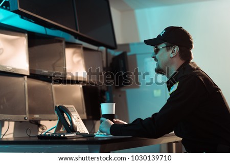 side view of smiling prison guard sitting with cup of coffee and monitoring people in jail
