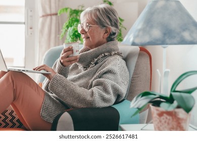 Side view of smiling senior Caucasian woman sitting comfortably in armchair using laptop computer on her knees while have a coffee cup - Powered by Shutterstock