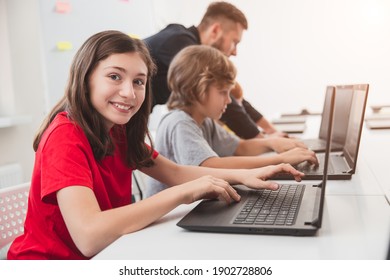 Side View Of Smiling Teen Secondary School Girl Looking At Camera While Using Laptop During Computer Science Lesson In Classroom With Teacher And Classmates