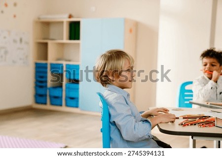 Side view of a smiling and thoughtful blonde school-aged European boy in a blue shirt and a dark-haired Black school-aged boy sitting at a desk in a school class.