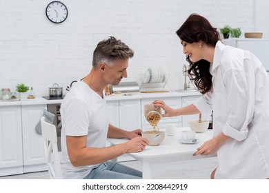 Side View Of Smiling Woman In Shirt Pouring Cereal In Bowl Near Husband And Coffee In Kitchen