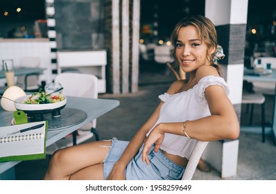 Side View Of Smiling Woman Sitting At Table On Chair In Outdoor Cafe With Open Notebook And Dish And Wearing White Blouse With Denim Skirt And Looking At Camera