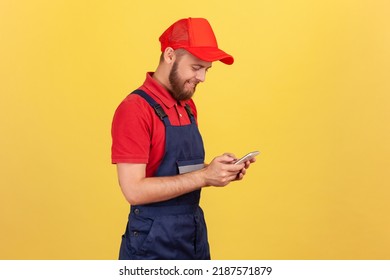 Side View Of Smiling Worker Man Typing On Mobile Phone, Using Cellphone Messenger To Accept Online Order As Delivery, Repair And Maintenance Services. Indoor Studio Shot Isolated On Yellow Background.