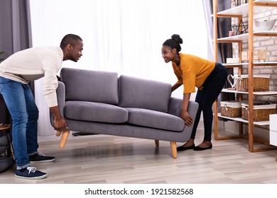 Side View Of A Smiling Young African Couple Lifting Sofa In Living Room