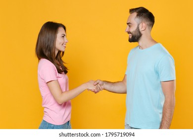 Side View Of Smiling Young Couple Friends Guy Girl In Blue Pink T-shirts Isolated On Yellow Wall Background Studio Portrait. People Lifestyle Concept. Mock Up Copy Space. Holding Greeting With Hands.